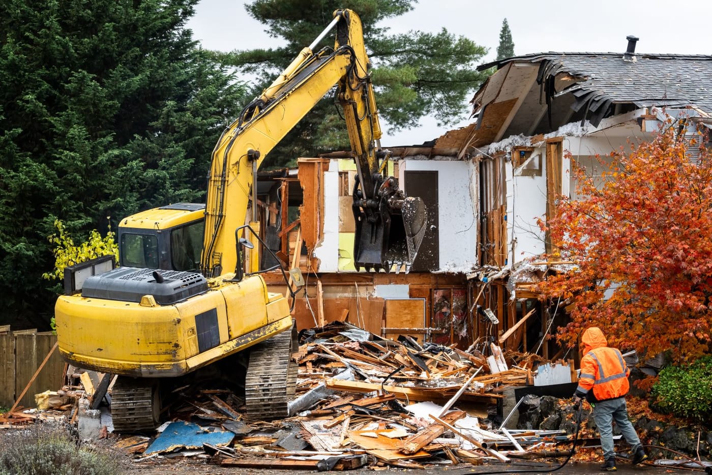 An excavator tearing down a house while an HS Dumpsters crew member manages the demolition site in New Jersey